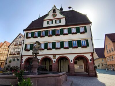 Ein historisches dreistöckiges Gebäude in Weil der Stadt mit grünen Fensterläden, Blumenkästen und einer Uhr an der Spitze; ein gewölbtes Erdgeschoss und eine Brunnenstatue stehen davor, Sonnenlicht scheint hinter dem Dach hervor.