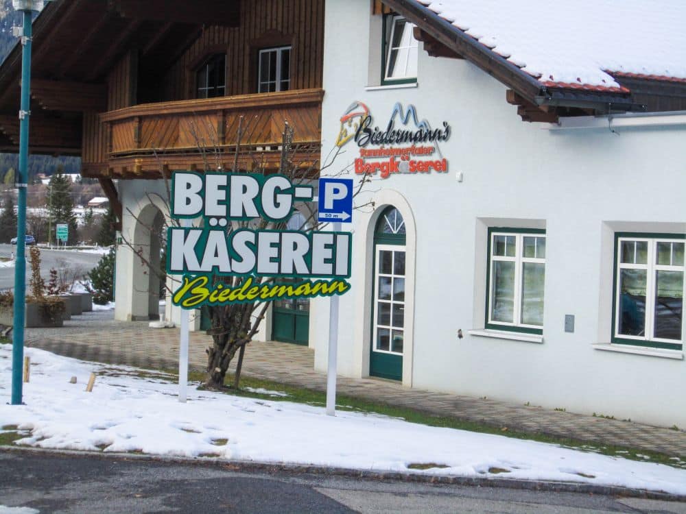 Ein Schild mit der Aufschrift Berg-Käserei Biedermann steht vor einem weißen Gebäude im Tannheimer Tal, dessen Boden und Dach mit Schnee bedeckt sind. Das Gebäude verfügt über einen Holzbalkon und trägt denselben Firmennamen an der Wand.