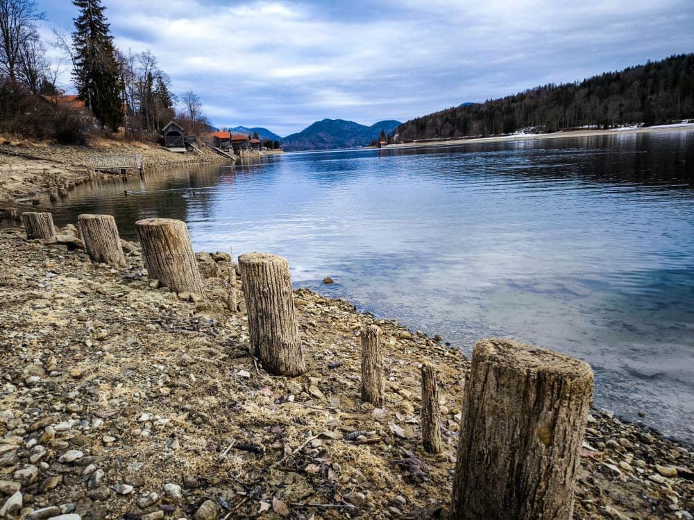 Verwitterte Holzpfähle säumen ein felsiges, abfallendes Ufer am ruhigen Walchensee, im Hintergrund sind baumbewachsene Hügel und verstreute Häuser unter einem wolkenverhangenen Himmel zu sehen.
