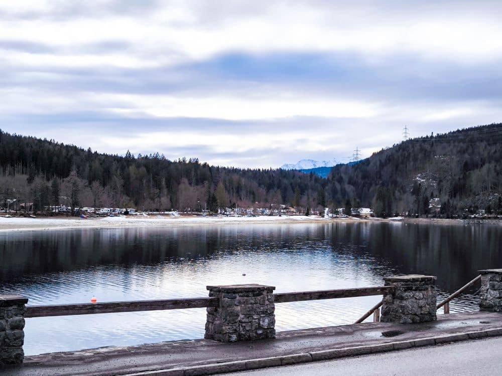 Ein ruhiger See mit Spiegelungen von Bäumen und bewölktem Himmel, umgeben von bewaldeten Hügeln in der Nähe des Walchensees. Ein Geländer aus Stein und Holz säumt den Straßenrand im Vordergrund, in der Ferne sind schneebedeckte Berge zu sehen.