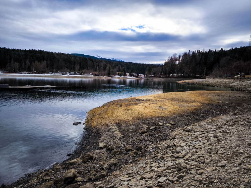 Ein felsiges Walchenseeufer mit klarem, ruhigem Wasser unter einem bewölkten Himmel. Kiefern und schneebedeckte Berge in der Ferne rahmen den Hintergrund ein, und einige Sonnenstrahlen beleuchten das Seeufer.