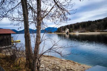 Ein ruhiger See, gesäumt von Bäumen mit kahlen Ästen, links ein hölzernes Bootshaus mit rotem Dach und in der Ferne Berge unter einem wolkenverhangenen Himmel. Auf der anderen Seite des ruhigen Walchensees kommen ein bewaldetes Ufer und ein kleiner Steg in Sicht.