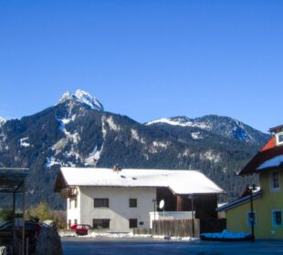 Ein weißes Haus mit einem schneebedeckten Dach steht vor einer Bergkette unter einem klaren blauen Himmel im Tannheimer Tal. Im Vordergrund sind weitere bunte Gebäude und Schneeflecken zu sehen.
