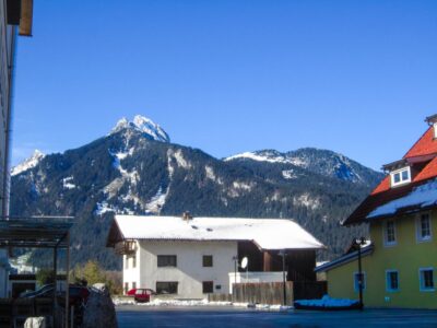 Ein weißes Haus mit einem schneebedeckten Dach steht vor einer Bergkette unter einem klaren blauen Himmel im Tannheimer Tal. Im Vordergrund sind weitere bunte Gebäude und Schneeflecken zu sehen.