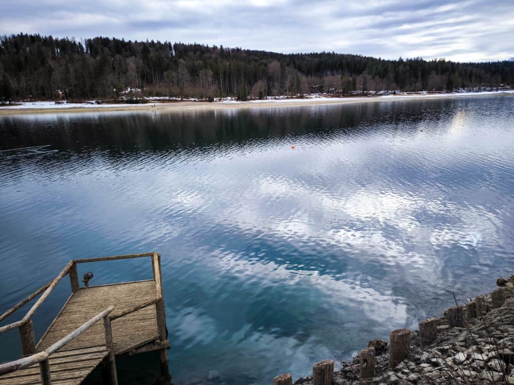 Ein hölzerner Steg erstreckt sich über das klare, ruhige Wasser des Walchensees, in dessen Wellen sich der bewölkte Himmel spiegelt. Bewaldete Hügel und Schneeflecken säumen das ferne Ufer im Hintergrund.