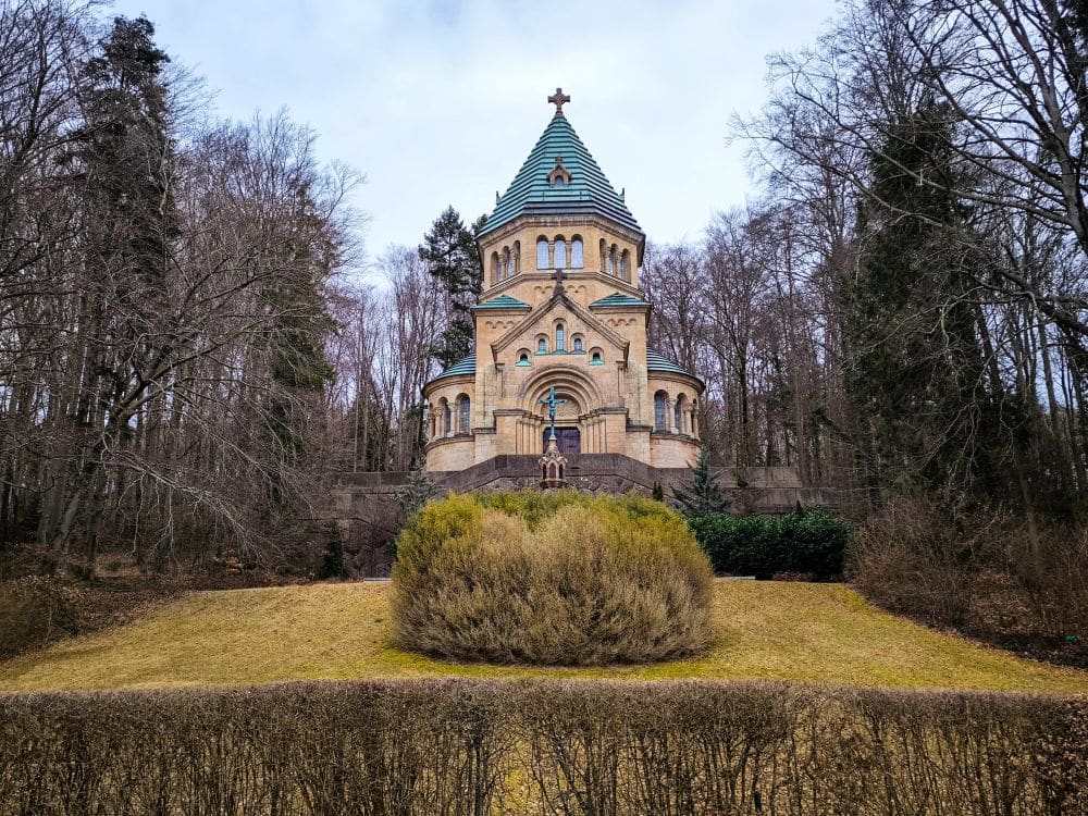 Eine historische Steinkapelle mit grünem Ziegeldach und Kreuz steht auf einer Anhöhe in der Nähe des Starnberger Sees, umgeben von blattlosen Bäumen und Sträuchern an einem bewölkten Tag, mit einem grasbewachsenen Vordergrund, der zum Gebäude hinaufführt.