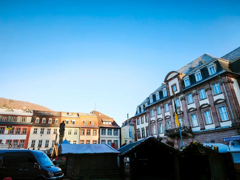 Bunte Gebäude im europäischen Stil und ein großes, verziertes Gebäude säumen einen offenen Platz in Heidelberg unter blauem Himmel. Im Vordergrund sind ein paar Zelte und ein dunkler Lieferwagen zu sehen.