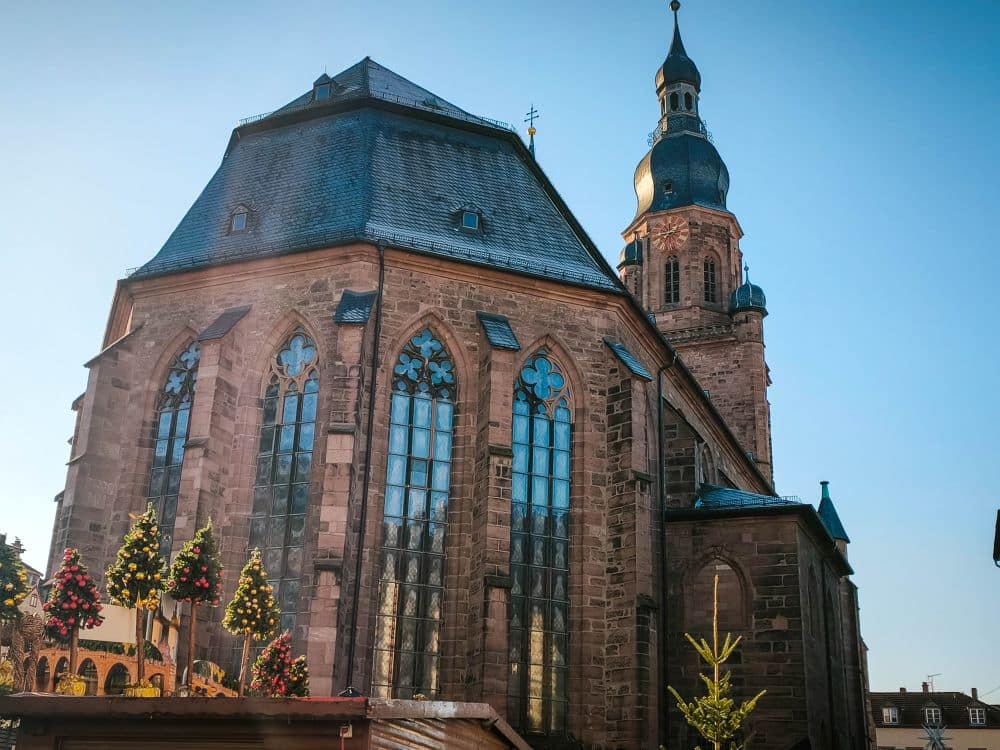 Eine große historische Kirche in Heidelberg mit hohen gewölbten Buntglasfenstern und einem Glockenturm vor einem klaren blauen Himmel. Geschmückte Weihnachtsbäume und festliche Marktstände verleihen dem lebendigen Vordergrund Charme.