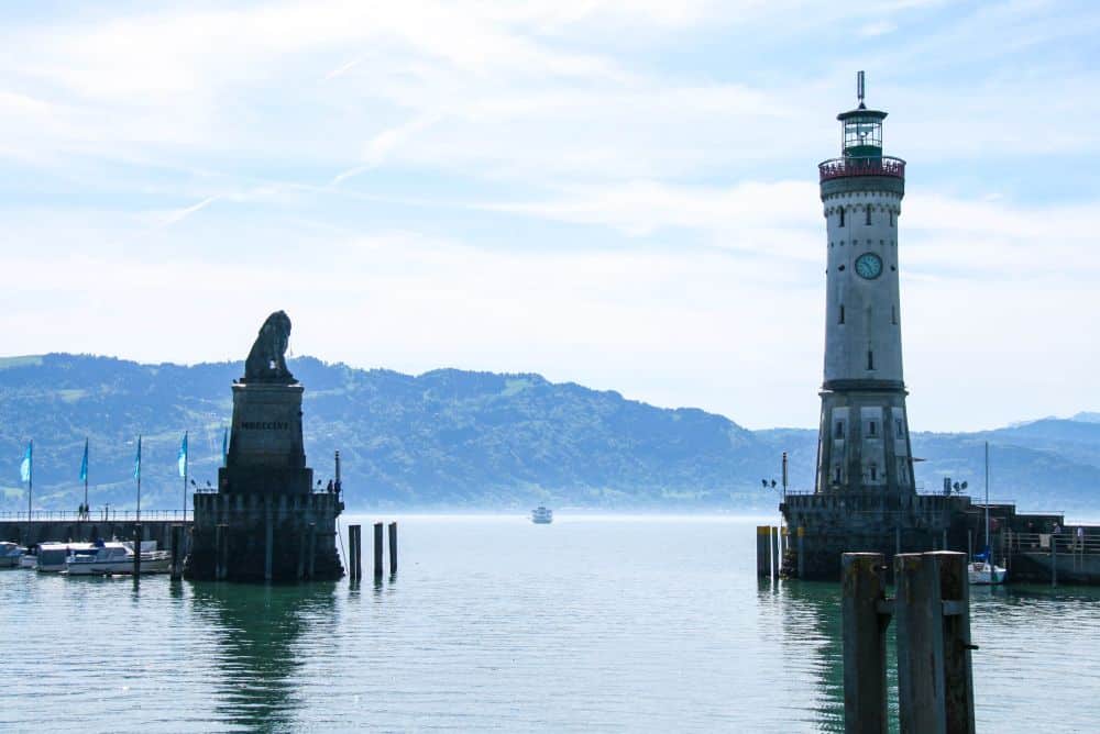 Eine malerische Hafeneinfahrt in Lindau am Bodensee, mit einer steinernen Löwenstatue auf der linken Seite und einem hohen weißen Leuchtturm mit Uhr auf der rechten Seite, mit Blick auf die Berge und ein Boot, das wahres Inselglück einfängt.