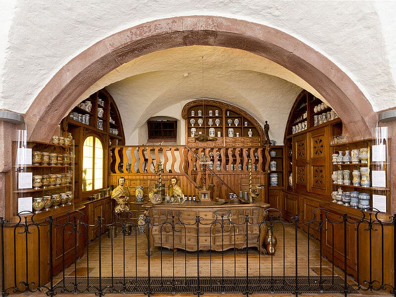 Eine historische Heidelberger Apotheke mit Holzregalen, die mit kunstvollen Gläsern und Flaschen bestückt sind, einem geschnitzten Holztresen und dekorativen Statuen, alles unter einem großen steinernen Torbogen mit einem warmen, einladenden Ambiente.