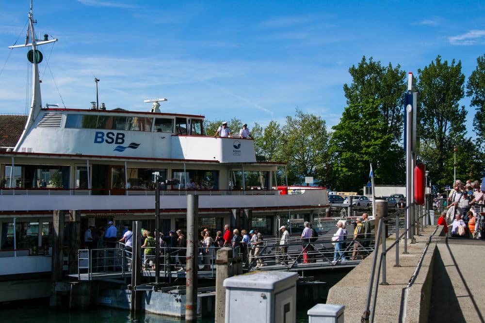 An einem sonnigen Tag in Lindau besteigt eine Menschenmenge eine BSB-Fähre an einer Anlegestelle. Im Hintergrund sind Bäume und Fahrzeuge zu sehen, bereit, das Inselglück am schönen Bodensee zu erleben.