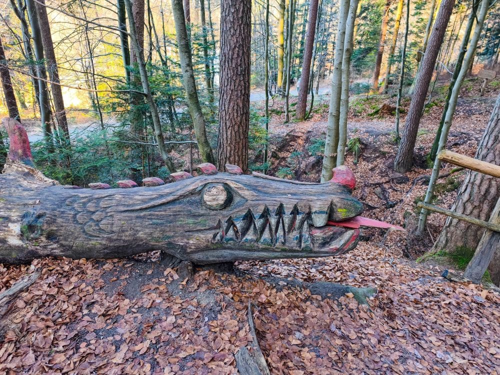 Ein großer Holzklotz, geschnitzt und bemalt wie ein Drachenkopf mit scharfen Zähnen, offenem Maul und roter Zunge, steht im Wald bei Herrenbach-Stausee, umgeben von hohen Bäumen und heruntergefallenen Blättern auf dem Boden.