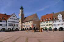 Ein großzügiger Stadtplatz mit Kopfsteinpflaster in Freudenstadt, umgeben von farbenfrohen traditionellen Gebäuden und einem hohen Uhrenturm unter einem klaren blauen Himmel. Nur wenige Menschen flanieren durch dieses Kleinod des Schwarzwalds, die Stadt der 1.000 Sonnenstrahlen.