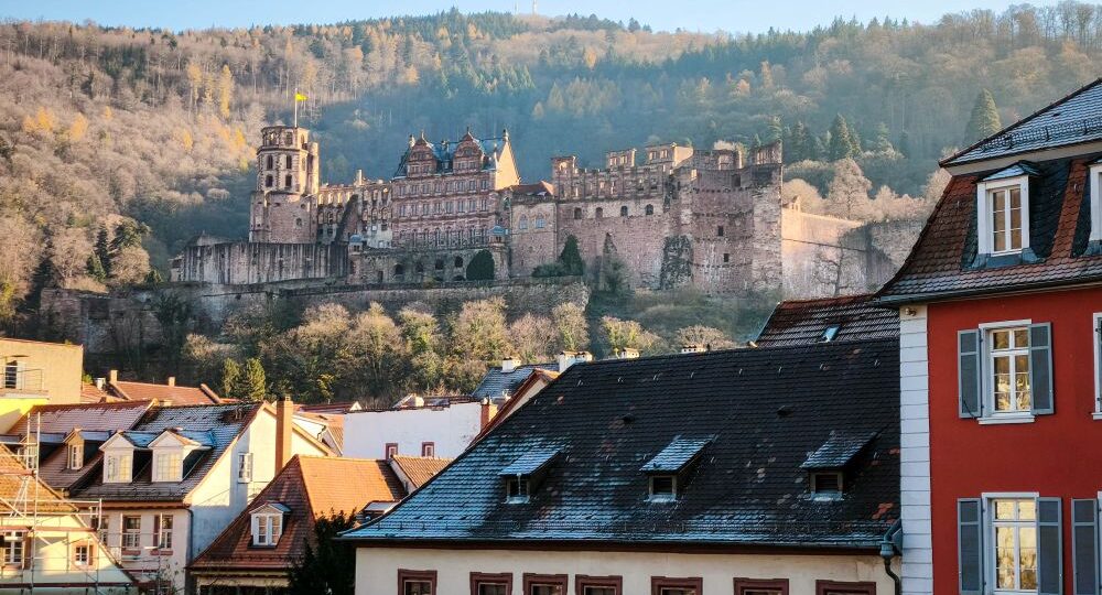 Ein großes historisches Schloss steht auf einem bewaldeten Hügel oberhalb von Heidelberg und ist in Sonnenlicht getaucht. Im Vordergrund sind Dächer und Gebäude der Stadt zu sehen, während der majestätische Hügel und das Heidelberger Schloss den Hintergrund dominieren.