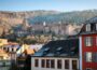 Ein großes historisches Schloss steht auf einem bewaldeten Hügel oberhalb von Heidelberg und ist in Sonnenlicht getaucht. Im Vordergrund sind Dächer und Gebäude der Stadt zu sehen, während der majestätische Hügel und das Heidelberger Schloss den Hintergrund dominieren.