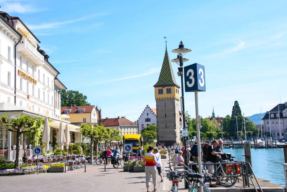 Eine belebte Uferpromenade in Lindau am Bodensee, mit Spaziergängern und Radfahrern, Straßencafés, abgestellten Fahrrädern und historischen Gebäuden. Der Turm mit dem gestreiften Dach und ein blaues Schild mit der Zahl 33 ergänzen das Inselglück bei strahlend blauem Himmel.