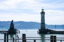 Eine malerische Hafeneinfahrt am Bodensee in Lindau mit einem hohen Leuchtturm auf der rechten und einer Löwenstatue auf der linken Seite, mit ruhigem Wasser, Segelbooten und fernen grünen Hügeln unter einem teilweise bewölkten Himmel - Inselglück pur.
