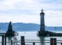 Eine malerische Hafeneinfahrt am Bodensee in Lindau mit einem hohen Leuchtturm auf der rechten und einer Löwenstatue auf der linken Seite, mit ruhigem Wasser, Segelbooten und fernen grünen Hügeln unter einem teilweise bewölkten Himmel - Inselglück pur.