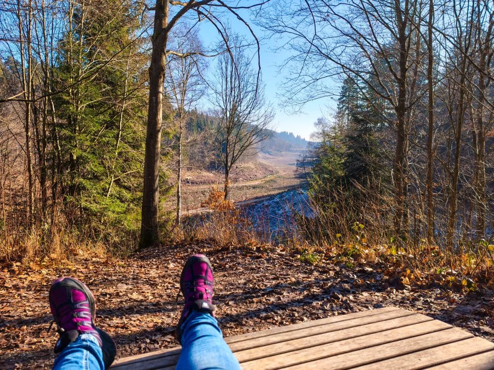 Eine Person in Jeans und lilafarbenen Turnschuhen, die sich auf einer Holzbank mit ausgestreckten Beinen ausruht, blickt auf das malerische Herrenbach-Stausee-Tal mit blattlosen Bäumen und einem klaren blauen Himmel an einem sonnigen Tag.