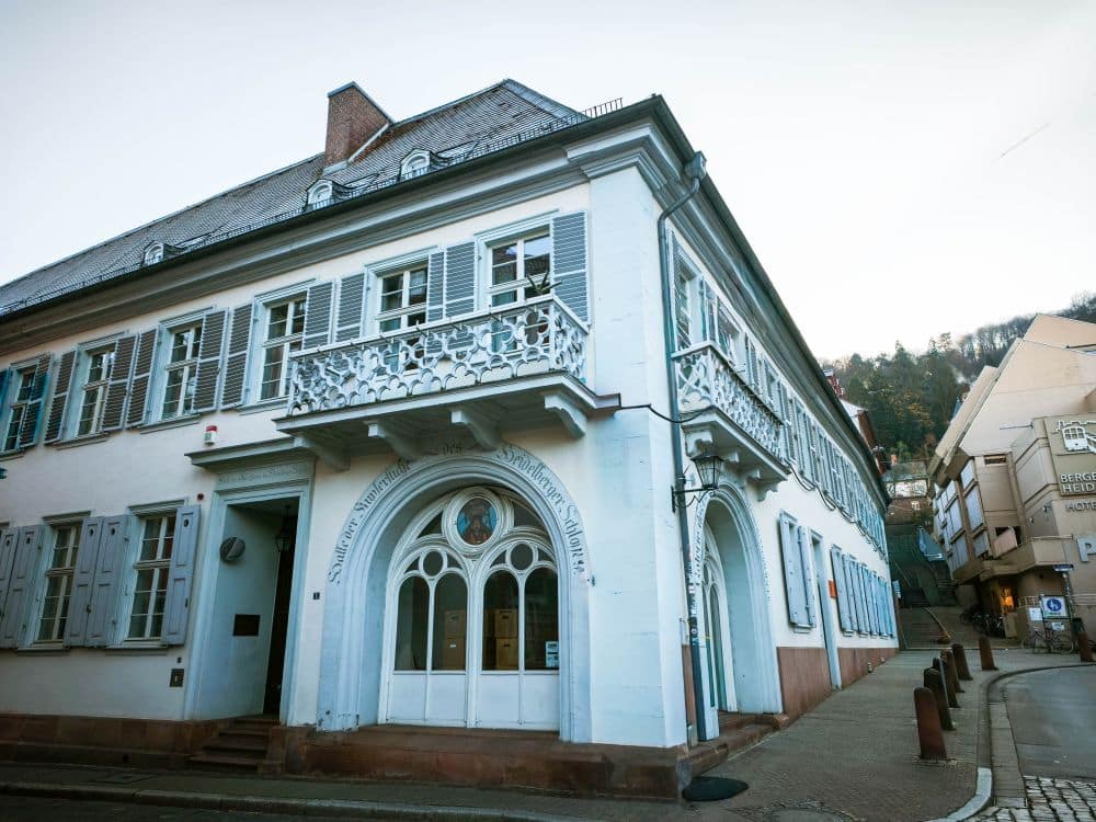 Ein historisches weißes Gebäude mit blauen Fensterläden und einem verzierten Balkon an einer ruhigen Heidelberger Straßenecke. Der gewölbte Eingang ist mit Zierleisten und einem runden Fenster über der Tür versehen. Im Hintergrund sind andere Gebäude und ein Hügel zu sehen.