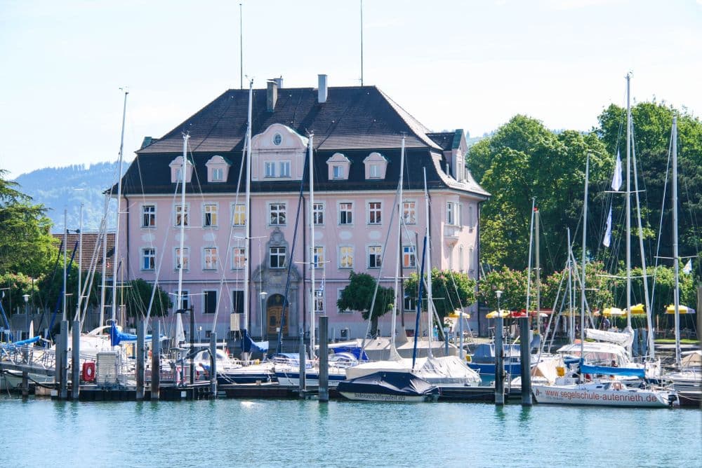 Ein großes rosafarbenes Gebäude mit einem dunklen Dach steht hinter einem Yachthafen voller Segelboote und Yachten in Lindau am Bodensee, mit Bäumen und Hügeln im Hintergrund - eine perfekte Szene des Inselglücks an einem sonnigen Tag.