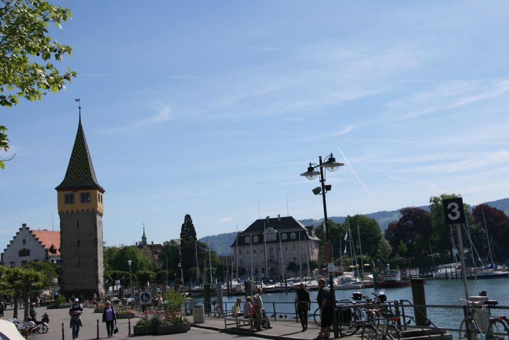 Eine Wasserszene in Lindau am Bodensee, mit spazierenden Menschen, abgestellten Fahrrädern, einem hohen historischen Turm mit Spitzdach, Booten im Yachthafen und Gebäuden und Hügeln im Hintergrund unter einem klaren blauen Himmel - Inselglück pur.