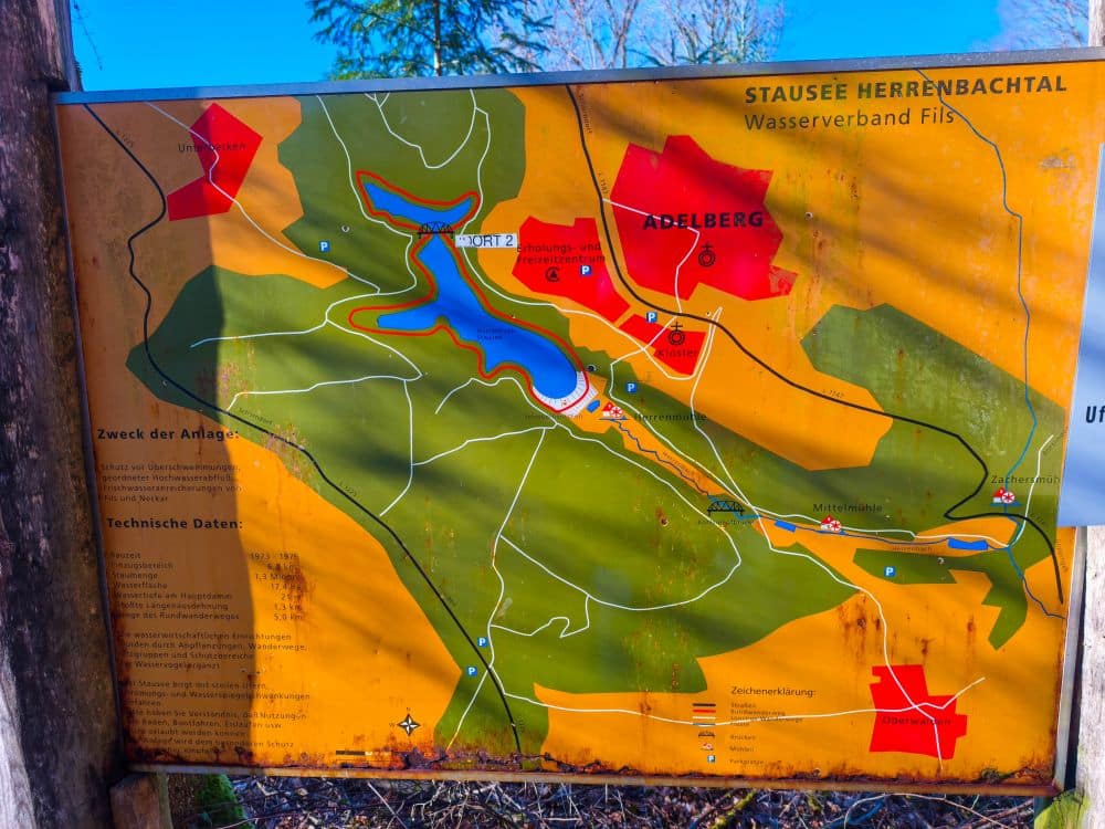 Eine verwitterte Karte zeigt das Gebiet des Herrenbach-Stausees im Schurwald mit Wanderwegen, Wasserspielen, nahegelegenen Orten und Naturinformationen in deutscher Sprache, umrahmt von Bäumen und Sonnenlicht.