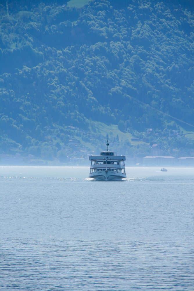 Ein weißes Fährboot fährt über das ruhige, blaue Wasser des Bodensees auf die Kamera zu. Im nebligen Hintergrund sind ein bewaldeter Berg und kleine Gebäude am Seeufer von Lindau zu sehen - echtes Inselglück.