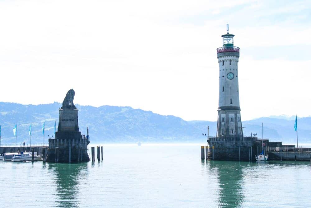 Eine steinerne Löwenstatue und ein hoher Leuchtturm stehen an der Hafeneinfahrt in Lindau. Unter dem wolkenverhangenen Himmel sind der ruhige Bodensee, die vertäuten Boote und die fernen Berge zu sehen.