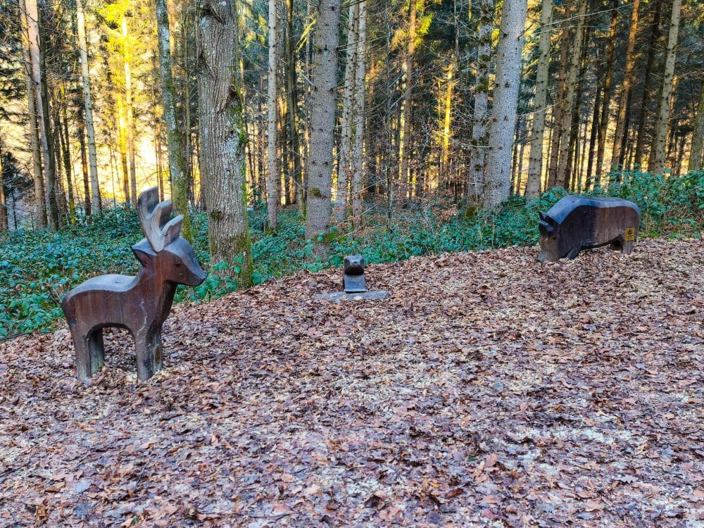 Die Holzskulpturen eines Rehs, eines Wildschweins und eines Eichhörnchens stehen auf einem mit trockenem Laub bedeckten Waldboden in der Nähe des Herrenbach-Stausees, umgeben von hohen Bäumen und üppigem Grün im Hintergrund.