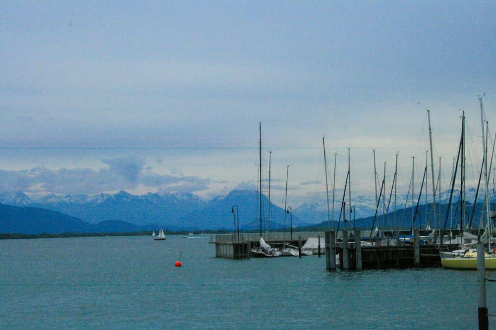 Segelboote im Yachthafen auf dem ruhigen blauen Wasser des Bodensees, mit schneebedeckten Bergen im Hintergrund unter einem bewölkten Himmel. Ein paar Segelboote und eine rote Boje treiben in der Nähe von Lindau und ergänzen diese Szene von Inselglück.
