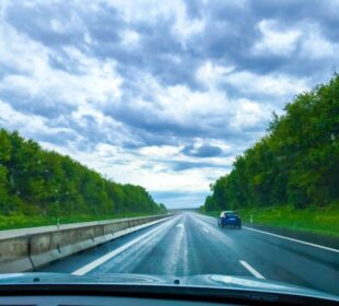 Blick aus einem Auto, das auf einer nassen, leeren, von grünen Bäumen gesäumten Landstraße unter bewölktem Himmel fährt; ein einzelnes Auto ist auf der rechten Spur zu sehen.