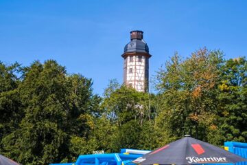 Ein hoher, traditioneller Fachwerkturm erhebt sich über grünen Bäumen auf dem Possen in Sondershausen, mit blauen aufblasbaren Abenteuerstrukturen und einem schwarzen Köstritzer-Schirm im Vordergrund unter einem klaren blauen Himmel.