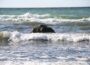 Ein großer Felsen liegt im flachen Meerwasser, während die Wellen unter einem klaren Himmel an ihm zerschellen - eine typische Szenerie in der Nähe der beliebten Ostseebäder. Das Wasser ist dunkelblau, mit weißem Schaum, der sich auf den brechenden Wellen bildet.