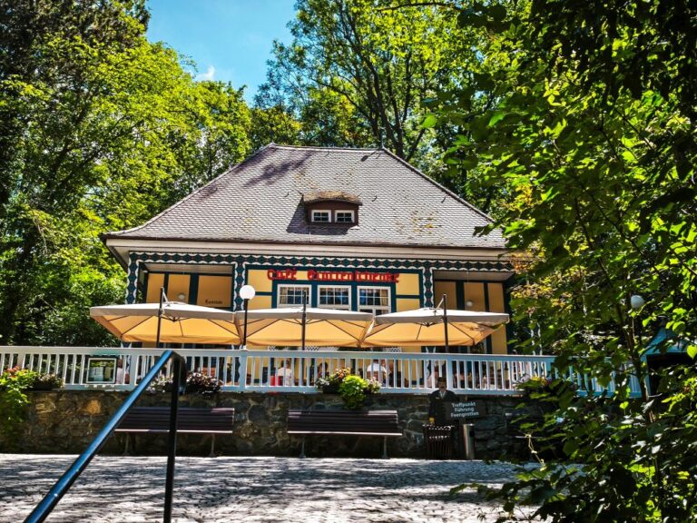 Saalfelder Feengrotten: Preise, Öffnungszeiten & Tipps für deinen Besuch 53 Ein charmantes Café mit schrägem Dach und Sitzgelegenheiten im Freien – komplett mit Sonnenschirmen und Blumenkästen – heißt die Gäste in der Nähe der Saalfelder Feengrotten willkommen. An einem sonnigen Tag, umgeben von Bäumen, ist es der perfekte Ort, um sich nach einem Besuch der nahegelegenen Sehenswürdigkeit zu entspannen; vergessen Sie nicht, sich vor Ihrem Besuch über Öffnungszeiten und Preise zu informieren.
